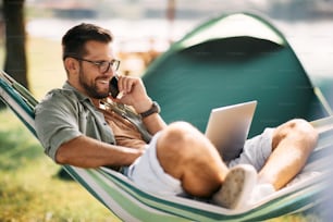 Imprenditore felice che si rilassa sull'amaca mentre utilizza il laptop e fa una telefonata durante la giornata in campeggio.