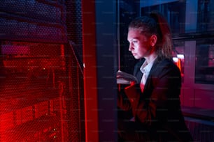 Female administrator using notebook computer while analyzing server in server room of data center lit by red light