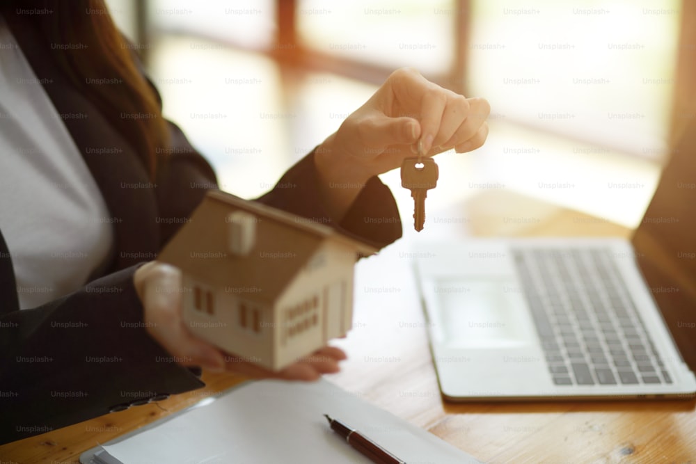 Cropped image of female Real Estate Agent broker holding home keychain and house model in her hand, sits at the desk in office.