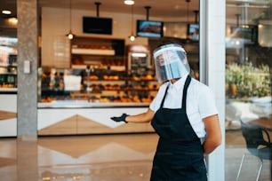 Schöne Frau mit Gesichtsschutz und Schutzmaske, die in einer Bäckerei oder einem Fast-Food-Restaurant arbeitet. Sie steht vor dem Laden und hält mit verschränkten Armen Desinfektionsmittel in der Hand. Covid-19-Sicherheitsmaßnahmen.