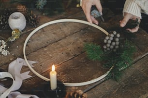 Making modern minimalistic christmas wreath. Hands making wreath on rustic table with fir branches, brunia herb, ribbons, round wooden hoop, thread, candle. Atmospheric moody image