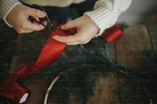 Hände im kuscheligen Pullover schneiden rotes Band mit Schere für modernen traditionellen Kranz mit Tannenzweigen und Holzreifen auf rustikalem Tisch. Atmosphärisches stimmungsvolles Bild. Stilvollen Weihnachtskranz basteln