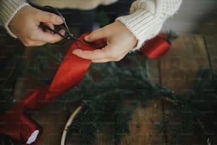 Hands in cozy sweater cutting red ribbon with scissors for modern traditional wreath with fir branches and wooden hoop on rustic table. Atmospheric moody image. Making stylish christmas wreath