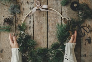 Hands holding stylish modern christmas wreath on rustic wooden table with branches, brunia, candle, scissors, thread, pine cones. Merry Christmas! Winter holidays preparation, atmospheric image