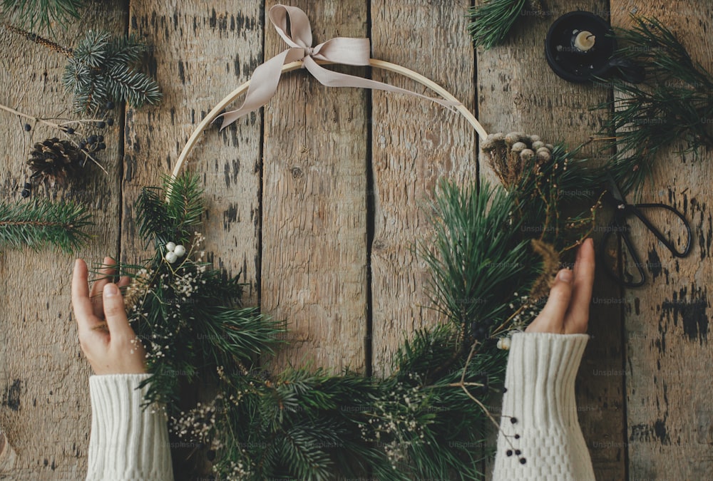 Hände mit stilvollem modernem Weihnachtskranz auf rustikalem Holztisch mit Zweigen, Brunia, Kerze, Schere, Faden, Tannenzapfen. Frohe Weihnachten! Winterurlaubsvorbereitung, stimmungsvolles Bild