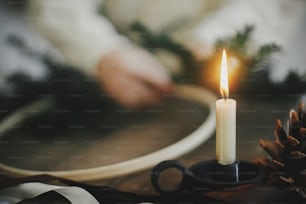 Stylish burning candle and pine cone on background of hands making modern christmas wreath with fir branches on rustic wooden table. Atmospheric image. Xmas advent. Winter holiday preparation