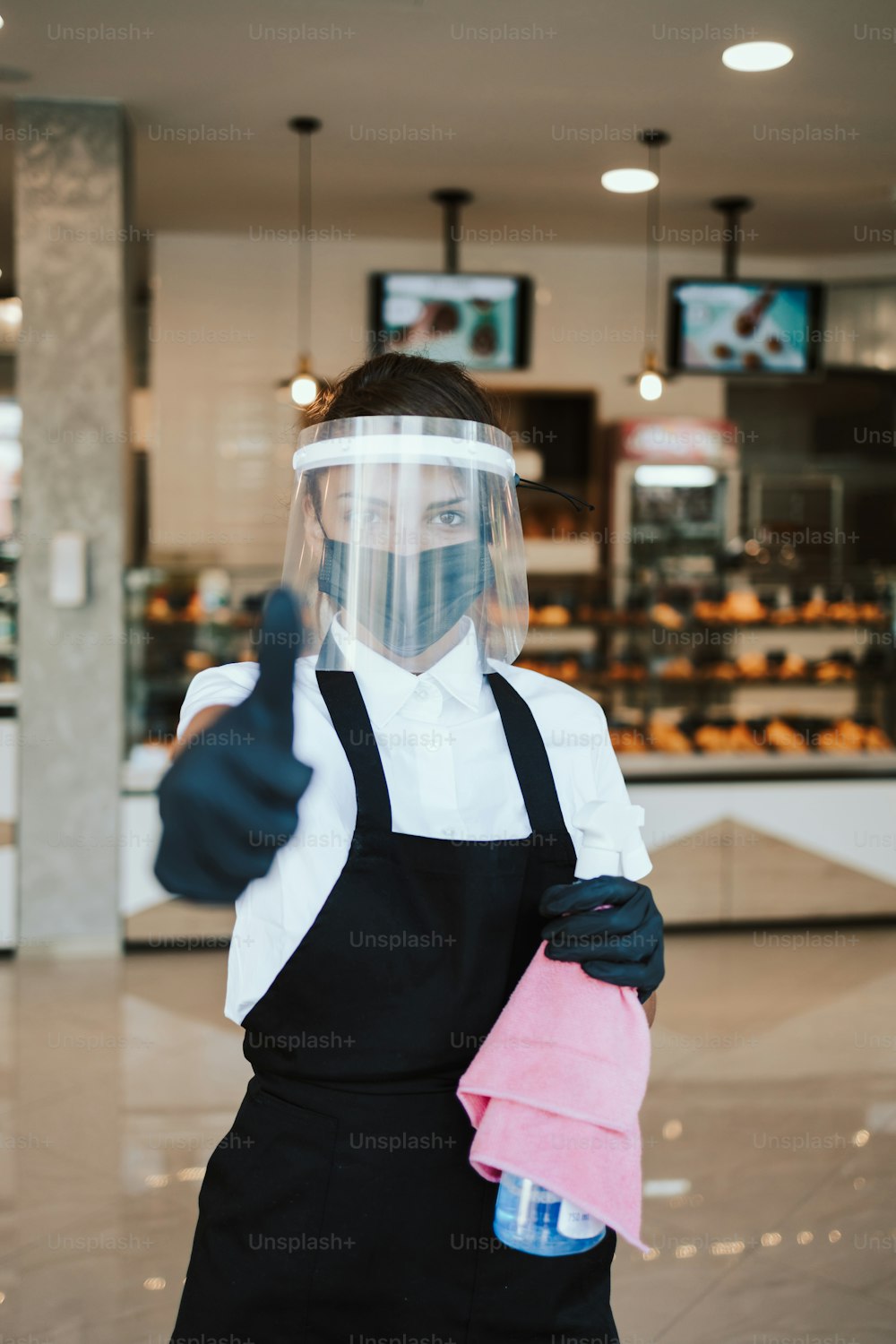 Beautiful woman with face shield and protective mask working bakery or fast food restaurant. She is standing in front of shop and holding disinfectants with arms crossed. Covid-19 safety measures.