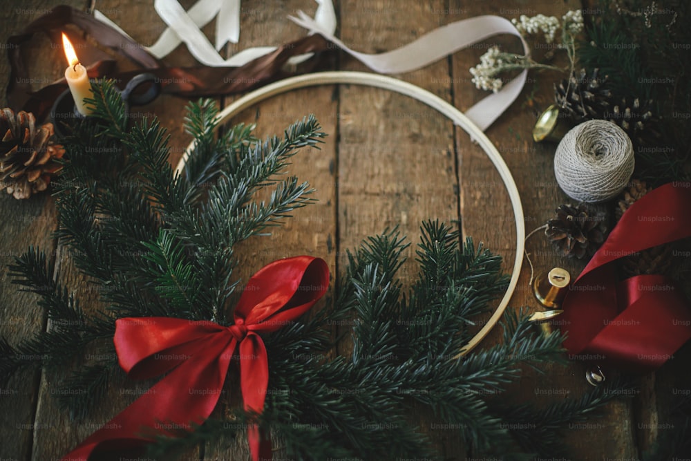 Buon Natale e Buone Feste! Ghirlanda natalizia moderna con rami di abete e fiocco rosso su tavolo rustico in legno con candela, nastri, pigne. Immagine atmosferica lunatica.