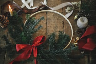 Merry Christmas and Happy holidays! Modern christmas wreath with fir branches and red bow on rustic wooden table with candle, ribbons, pine cones. Atmospheric moody image.