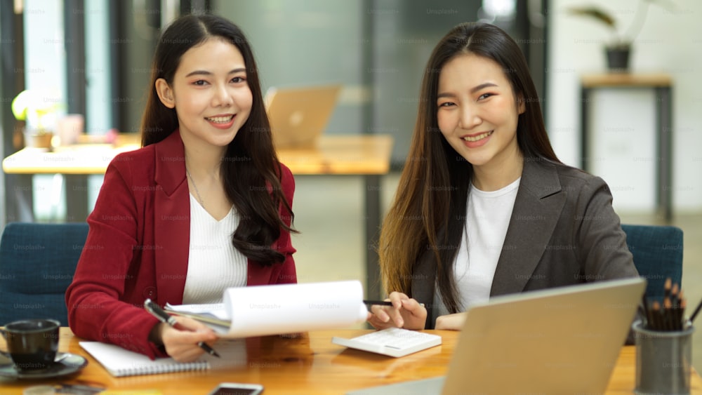 Fröhliche und freundliche attraktive asiatische Geschäftsfrauen oder Buchhalter arbeiten an ihrem Schreibtisch zusammen.