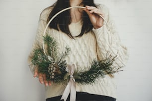 Mujer elegante con suéter acogedor que sostiene una corona navideña moderna sobre fondo blanco en el interior. Imagen atmosférica. Preparación y decoración de las vacaciones de invierno. Hacer corona navideña boho