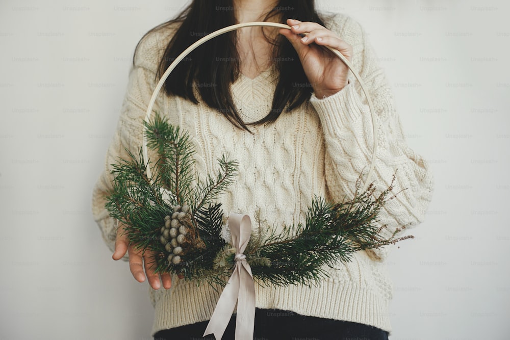 Stylish woman in cozy sweater holding modern christmas wreath on white background indoors. Atmospheric image. Winter holiday preparation and decor. Making boho christmas wreath