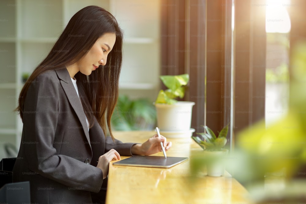 캐주얼 정장을 입은 매력적인 아시아 사업가의 측면 모습은 휴대용 태블릿 컴퓨터에서 작업하는 현대적인 밝은 카페에 앉아 있습니다.