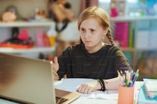 회색 셔츠를 입은 현대 소녀는 화창한 날에 집에서 원격 교육을 받는 노트북과 통합 문서를 가지고 있습니다.