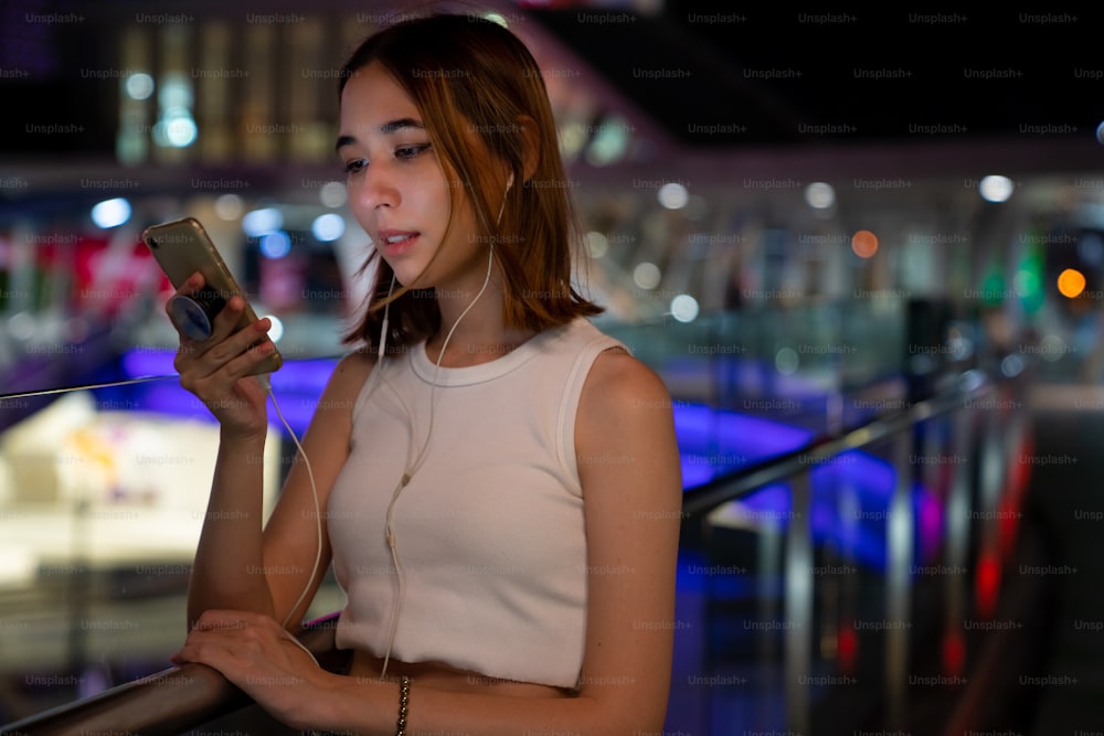 Schöne asiatische Frau, die die Stadtstraße entlang geht, die Musik von Kopfhörern auf Smartphone-Anwendung hört und die beleuchteten Nachtlichter der Stadt betrachtet. Hübsches Mädchen genießt urbanen Outdoor-Lifestyle in der Nacht