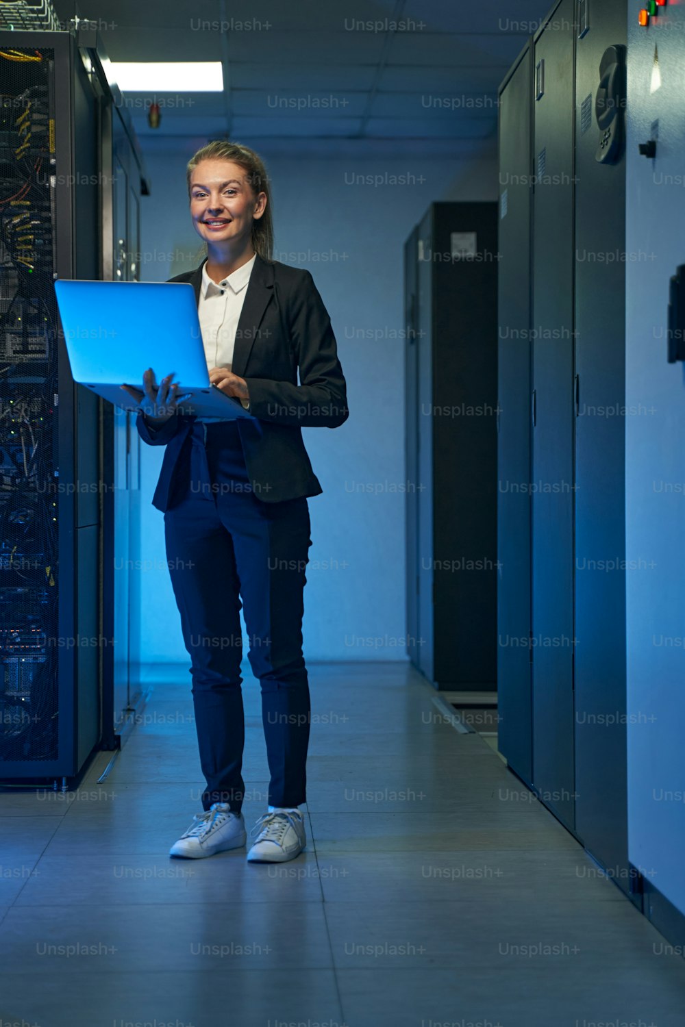 Happy female administrator in elegant suit working with laptop computer while standing between server racks of data center