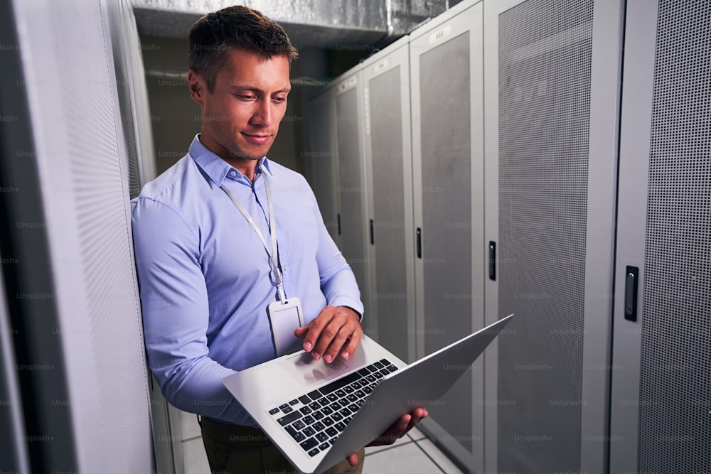 Concentrated Caucasian IT professional standing in server room and typing on laptop