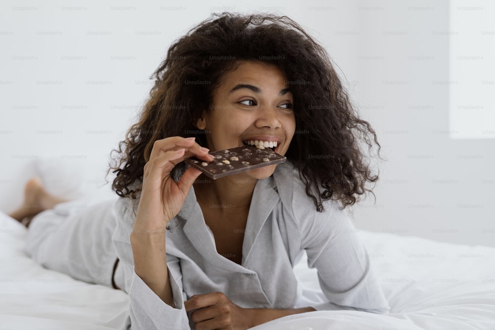 Concetto di cibo squisito. Donna afroamericana felice che mangia cioccolato svizzero marrone, sdraiata sul letto in pigiama e trascorre una mattinata luminosa a casa. Ragazza millenaria che gode di dessert dolce, guardando da parte