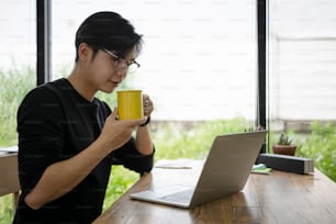 사려 깊은 청년 기업가는 뜨거운 커피를 마시고 노트북 컴퓨터에서 뉴스를 읽습니다.