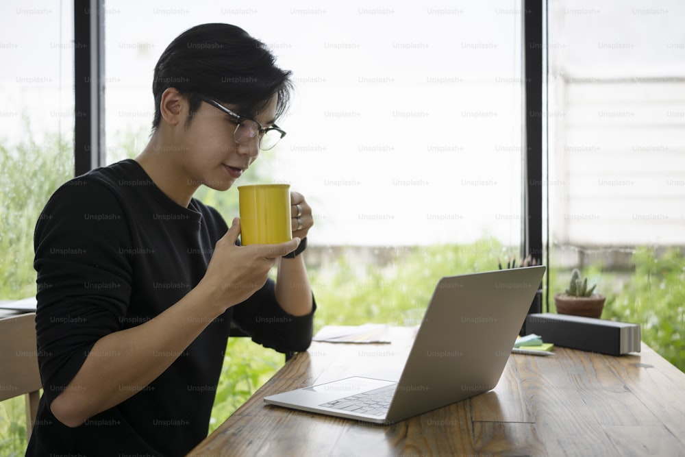 Jovem empreendedor pensativo bebendo café quente e lendo notícias em laptop.