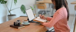 Side view of millennial female writing on her diary while looking at digital tablet computer, remotely working at cafe. tablet screen mockup