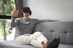 Young man wearing virtual reality headset playing video game or watching movie on sofa at home.