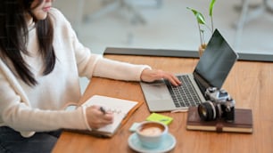 Cropped image, Millennial female freelance web blogger writing her online topics list in her book while working on laptop at coffee shop.