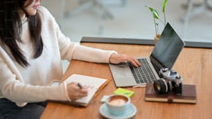 Image recadrée, blogueuse web indépendante de la génération Y écrivant sa liste de sujets en ligne dans son livre tout en travaillant sur un ordinateur portable dans un café.