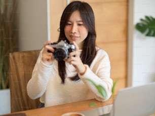 Charming asian young female photographer, content editor holding vintage retro camera, sits and works on her laptop at cafe.