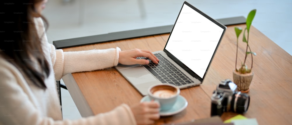 Primer plano de una fotógrafa independiente o bloguera de café seleccionando una fotografía, trabajando y escribiendo una reseña de café en su computadora portátil en un café.