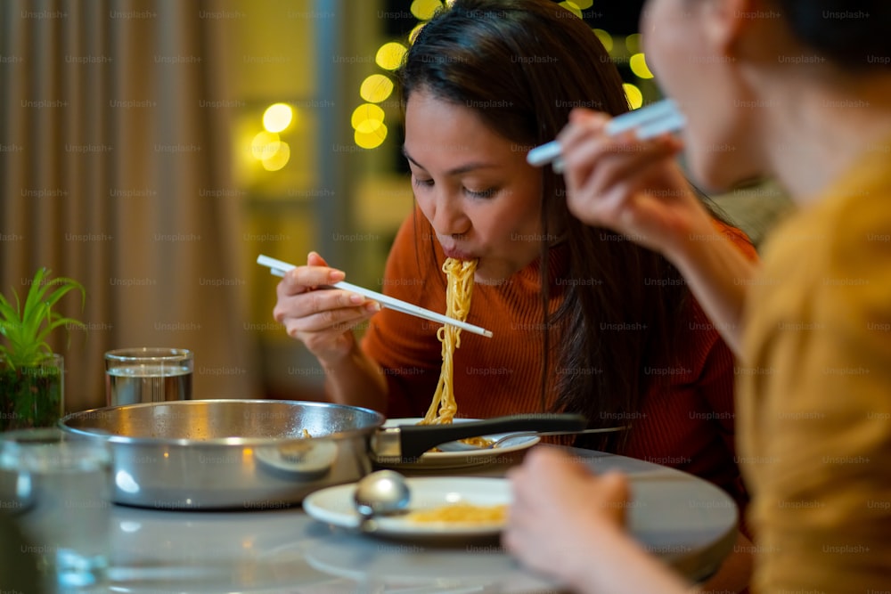 Beautiful Asian woman friends eating instant ramen noodle with sausage and talking together in living room at night. Female couple girlfriend enjoy indoor lifestyle having dinner together at home