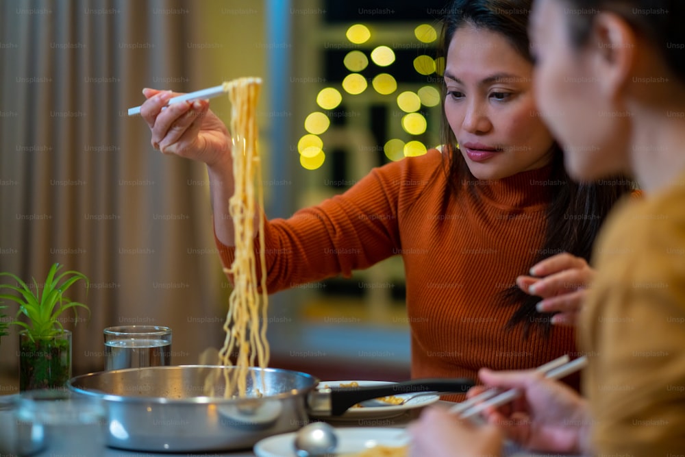 Schöne asiatische Freundinnen essen Instant-Ramen-Nudeln mit Wurst und unterhalten sich nachts im Wohnzimmer. Weibliches Paar Freundin genießt Indoor-Lifestyle beim gemeinsamen Abendessen zu Hause