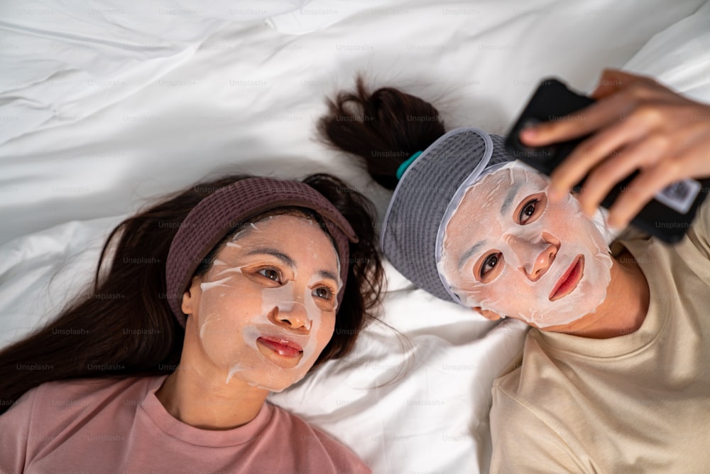 Smiling Asian woman friends sitting on the bed with applying skin care treatment facial mask on their face together at home. Female gay couple using smartphone taking selfie together with happiness