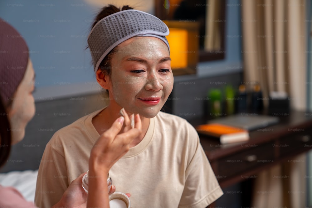 Amies asiatiques souriantes assises sur le lit avec l’application d’un masque facial de soin de la peau sur leur visage ensemble à la maison. Un couple gay féminin se détend et profite d’un traitement facial de beauté avec bonheur