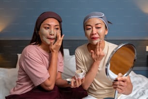Smiling Asian woman friends sitting on the bed with applying skin care facial mask on their face together at home. Female gay couple relax and enjoy beauty facial treatment together with happiness