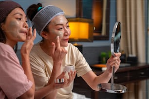 Smiling Asian woman friends sitting on the bed with applying skin care facial mask on their face together at home. Female gay couple relax and enjoy beauty facial treatment together with happiness