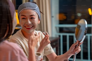 Smiling Asian woman friends sitting on the bed with applying skin care facial mask on their face together at home. Female gay couple relax and enjoy beauty facial treatment together with happiness