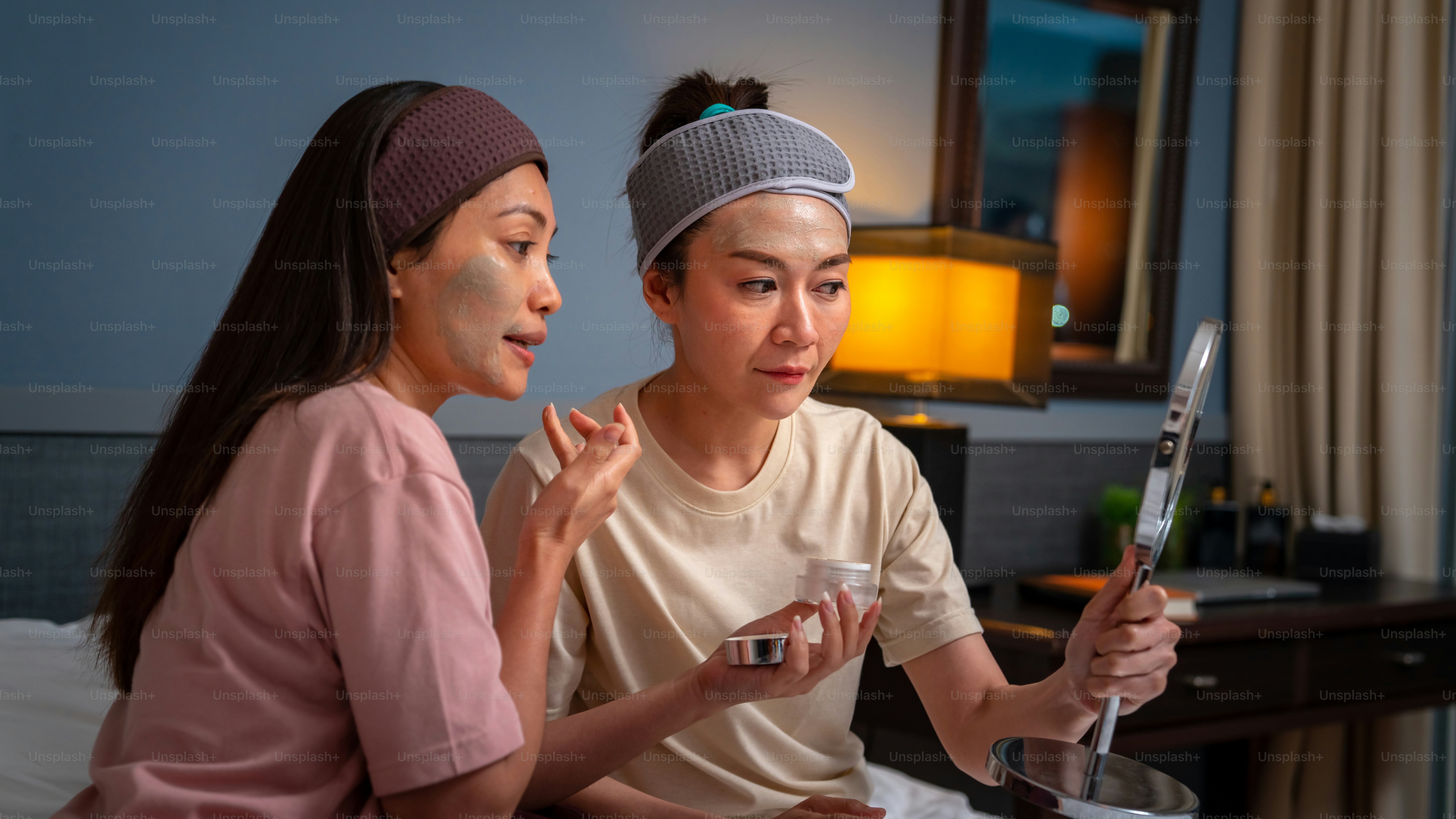 Smiling Asian woman friends sitting on the bed with applying skin care facial mask on their face together at home. Female gay couple relax and enjoy beauty facial treatment together with happiness