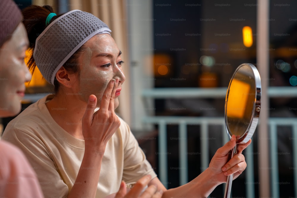 Amigas asiáticas sonrientes sentadas en la cama con la aplicación de una máscara facial para el cuidado de la piel en la cara juntas en casa. Pareja gay femenina se relaja y disfruta de un tratamiento facial de belleza junto con la felicidad
