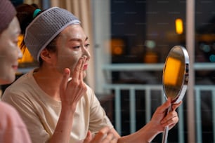 Smiling Asian woman friends sitting on the bed with applying skin care facial mask on their face together at home. Female gay couple relax and enjoy beauty facial treatment together with happiness