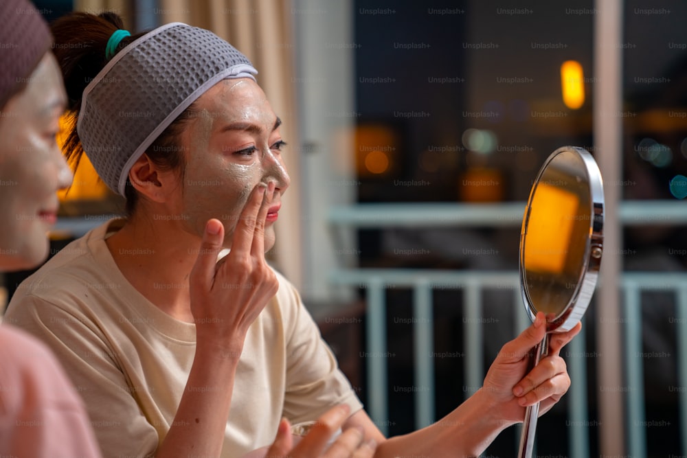 Smiling Asian woman friends sitting on the bed with applying skin care facial mask on their face together at home. Female gay couple relax and enjoy beauty facial treatment together with happiness