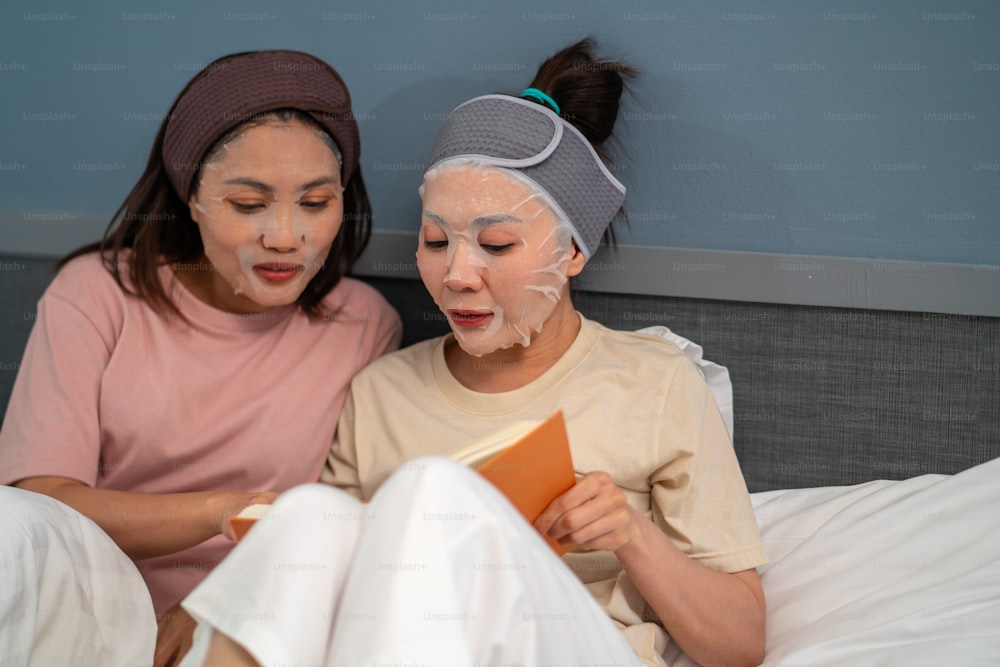 Amies asiatiques souriantes assises sur le lit avec l’application d’un masque facial de soin de la peau sur leur visage ensemble à la maison. Un couple gay féminin se détend et profite d’un soin du visage de beauté et de la lecture d’un livre ensemble.