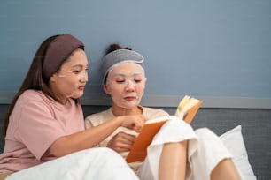 自宅で一緒に顔にスキンケアフェイシャルマスクを塗ってベッドに座って笑顔のアジア人女性の友人。女性のゲイカップルはリラックスして、美容フェイシャルトリートメントを楽しんだり、一緒に本を読んだりします。