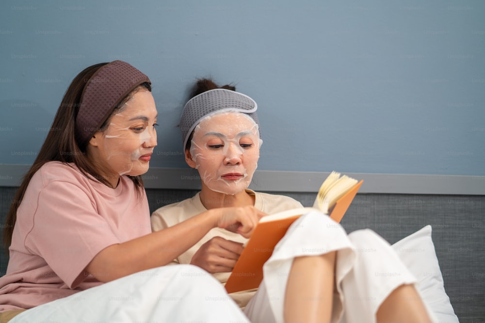 Amiche asiatiche sorridenti sedute sul letto con l'applicazione della maschera facciale per la cura della pelle sul viso insieme a casa. Coppia gay femminile rilassarsi e godere di un trattamento viso di bellezza e leggere un libro insieme.