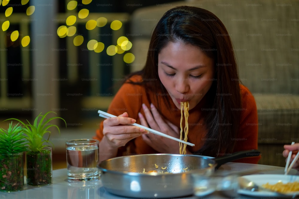 Belle amiche asiatiche che mangiano ramen istantaneo con salsiccia e parlano insieme in soggiorno di notte. La fidanzata di coppia femminile si gode lo stile di vita al coperto cenando insieme a casa