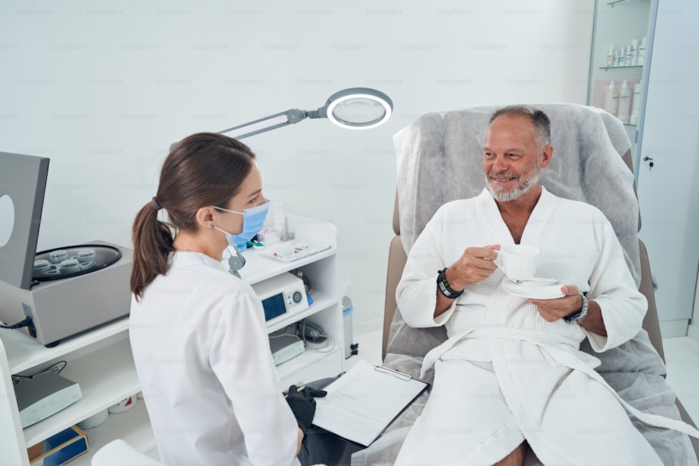Joyful male client in bathrobe drinking coffee and talking with beautician while woman in medical mask holding clipboard