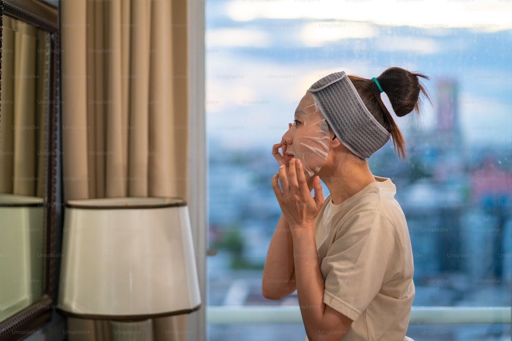 Asian woman standing in front of the mirror in bedroom and applying skin care facial face mask with serum on her face. Beautiful female enjoy self beauty facial spa and body skin treatment at home