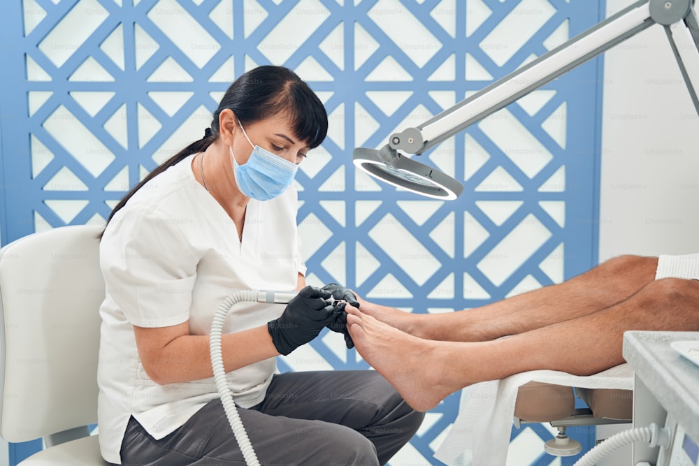 Maître de pédicure dans un masque médical à l’aide d’une machine électrique professionnelle tout en épluchant les ongles des clients masculins dans un salon de spa