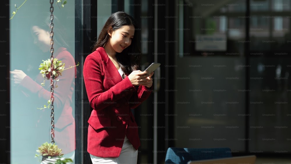 Businesswoman in red suit leaning on glass window while reading or texting message on smart mobile phone in office