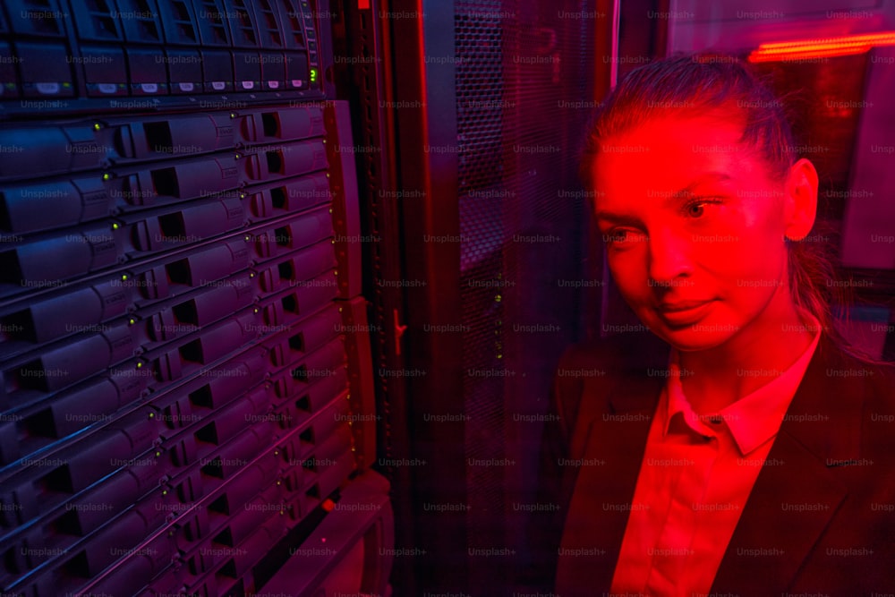Administratrice système attrayante regardant le disque dur d’un serveur rack dans un centre de données moderne, image de couleur rouge
